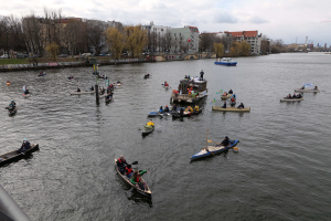 Eine Gruppe von Menschen in Kanus, die auf einem Fluss paddeln, während des Berliner Kanufestivals, mit einem Geländer auf der linken Seite, Bäumen, Gebäuden, Fahrzeugen und Pfählen im Hintergrund unter einem klaren blauen Himmel.