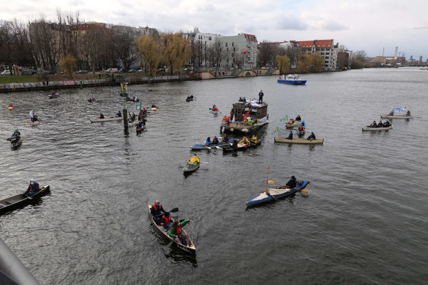 Eine Gruppe von Menschen in Kanus, die auf einem Fluss paddeln, während des Berliner Kanufestivals, mit einem Geländer auf der linken Seite, Bäumen, Gebäuden, Fahrzeugen und Pfählen im Hintergrund unter einem klaren blauen Himmel.