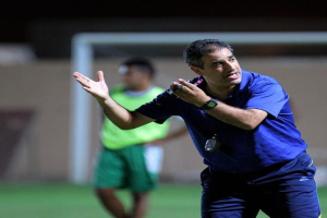 Ein Mann in einem blauen T-Shirt und Hose steht auf einem Fußballfeld und hält ein Mikrofon in der Hand, während er seinen Spielern Zeichen gibt. Im Hintergrund sind eine Person in einem grünen Kleid, ein Torpfosten und eine Wand zu sehen.