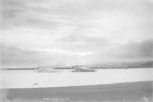 Ein Schwarz-Weiß-Foto eines Eisbergs, der im Ozean treibt, umgeben von Hügeln und einer bewölkten Himmel, mit dem Text "Sunset Childs Glacier" unten.