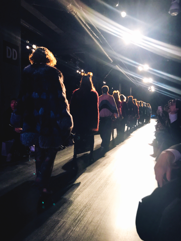 Eine Gruppe von Menschen läuft über den Laufsteg einer Fashion Show, während das Publikum auf beiden Seiten zuschaut.