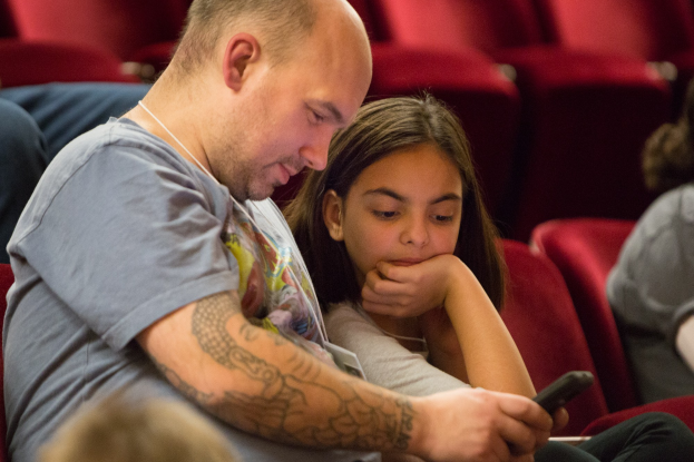 Ein Mann und ein junges Mädchen sitzen in einem Kino, beide schauen auf ein Handy, wobei der Mann es hält und das Mädchen konzentriert auf den Bildschirm schaut.