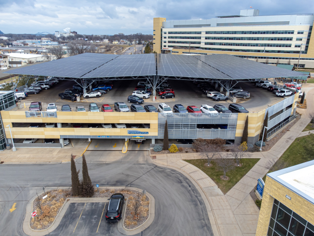 Luftaufnahme eines solarpanelbedeckten Parkplatzes mit parkenden Autos, umgeben von Gebäuden, Bäumen, Grüss, Straße und Hüllen unter einem bewölktem Himmel.