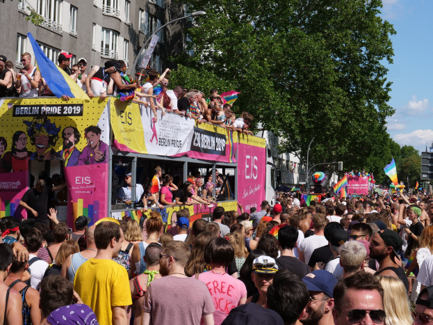 Eine große Menge marschiert während des Berlin Pride 2019 die Straße entlang, viele tragen Mützen und Schutzbrillen, einige halten Fahnen, mit Bannern, Laternenmasten, Bäumen und Gebäuden im Hintergrund unter einem bewölkten Himmel.