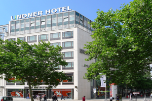 Großes gläsernes Hotelgebäude mit Text auf dem Dach, umgeben von Bäumen und einer belebten Straße mit Fußgängern und Fahrzeugen unter einem sichtbaren Himmel.