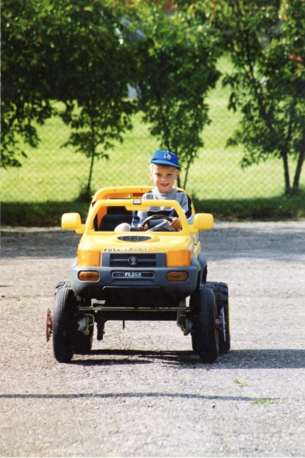 Ein junger Junge in einer Mütze fährt ein gelbes Spielzeugauto auf einer Straße, mit Bäumen und einem Maschendrahtzaun im Hintergrund.