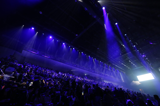 Eine große Menschenmenge sitzt vor einer Bühne mit Deckenbeleuchtung und ein Bildschirm ist auf der rechten Seite des Bildes zu sehen.