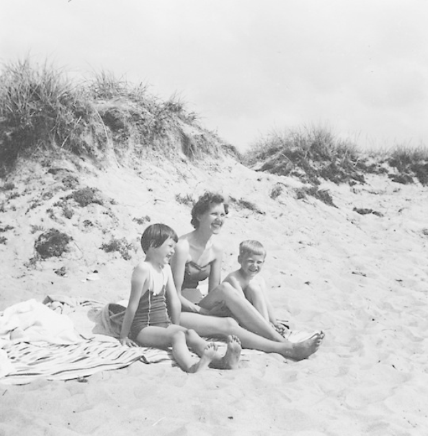 Schwarzes und weißes Foto einer Frau in einem Bikini und zwei Kindern in Badebekleidung, die auf einer Decke auf einem sandigen Strand mit Gras und Himmel im Hintergrund sitzen.