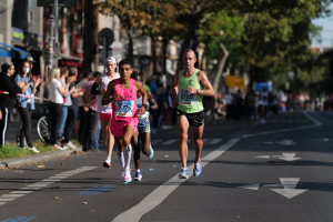 Eine Gruppe von Menschen, die bei einem Marathon laufen, auf einer Stadtstraße mit Zuschauern auf der linken Seite, unscharfen Rasen, Bäumen, Gebäuden, Pfählen, Brettern und einem Fahrrad im Hintergrund.