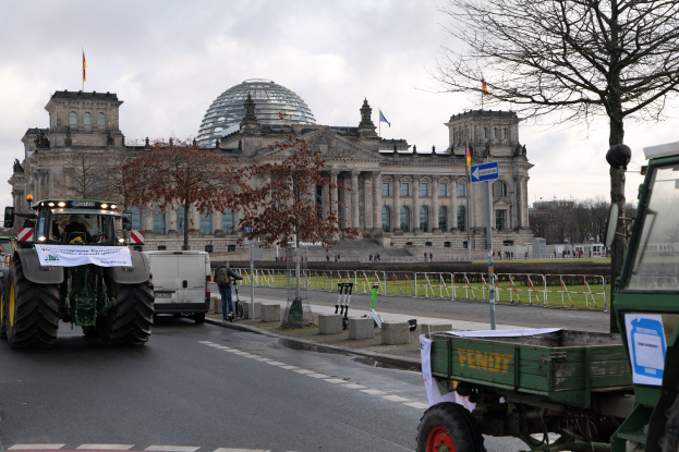 Eine Gruppe von Traktoren fährt vor dem Reichstaggebäude in Berlin, Deutschland, vorbei, das mit Fenstern, Säulen und Flaggen geschmückt ist und von Bäumen, Geländern, Pfählen und Brettern umgeben ist, mit ein paar Menschen im Hintergrund und einem bewölkten Himmel.