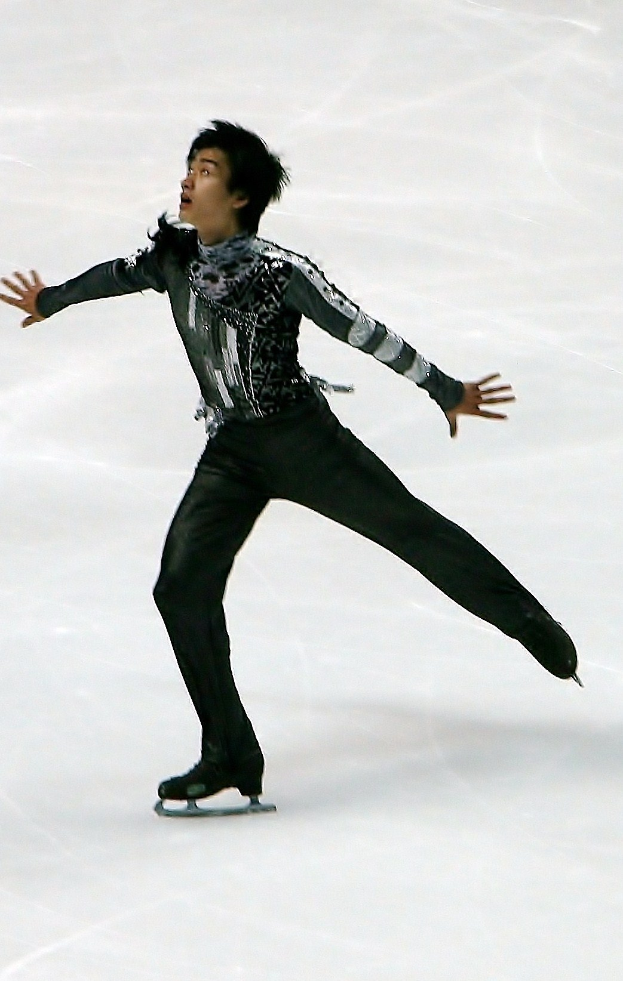 Männlicher Eiskunstläufer in schwarzer Kleidung gleitet über die Eisfläche und führt eine Choreografie mit synchronen Arm- und Beinbewegungen aus.