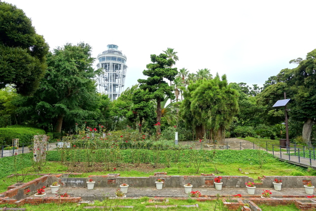 Ein Park mit einem Turm im Hintergrund, umgeben von grünem Gras, Pflanzen, Bäumen und verstreuten Blumentöpfen, mit einer Straße mit Geländern und einer am Mast angebrachten Tafel unter einem sichtbaren Himmel.