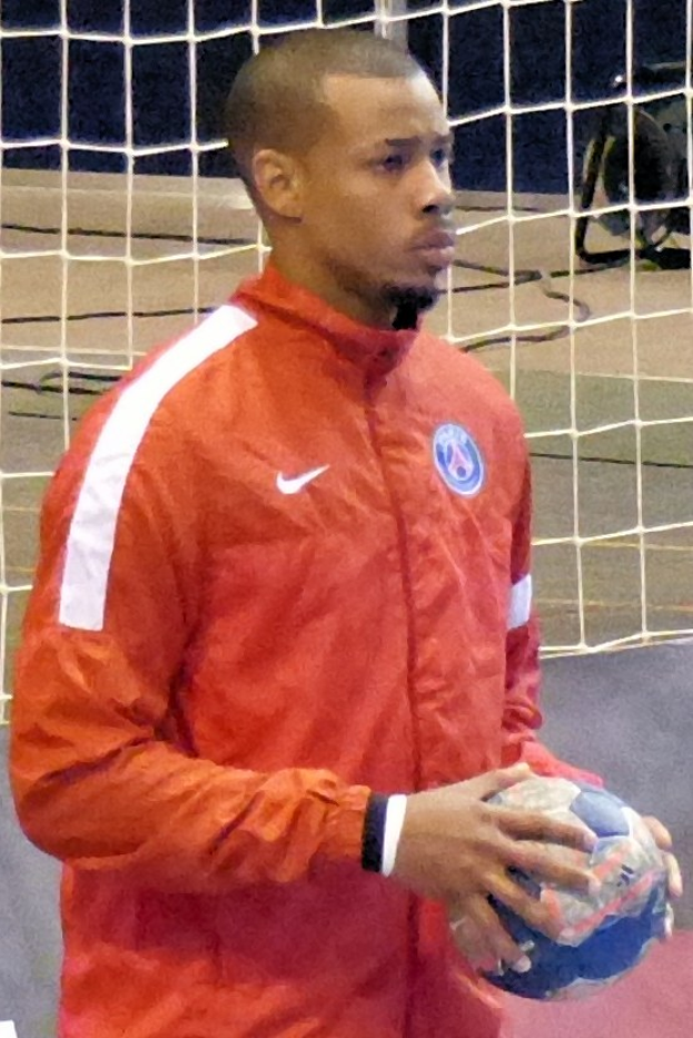 Ein Mann in einem blauen T-Shirt und Shorts steht vor einem Fußballtor und hält einen Fußball mit entschlossener Miene.