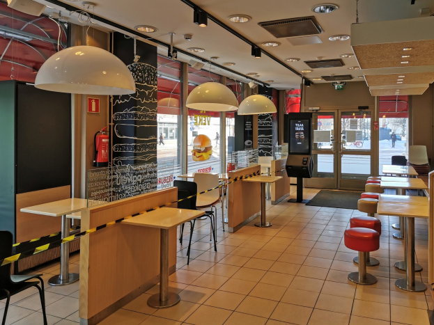Innenraum eines Schnellrestaurants mit Tischen, Stühlen, Hockern, Deckenleuchten, einem Feuerlöscher an der Wand und Glastüren.