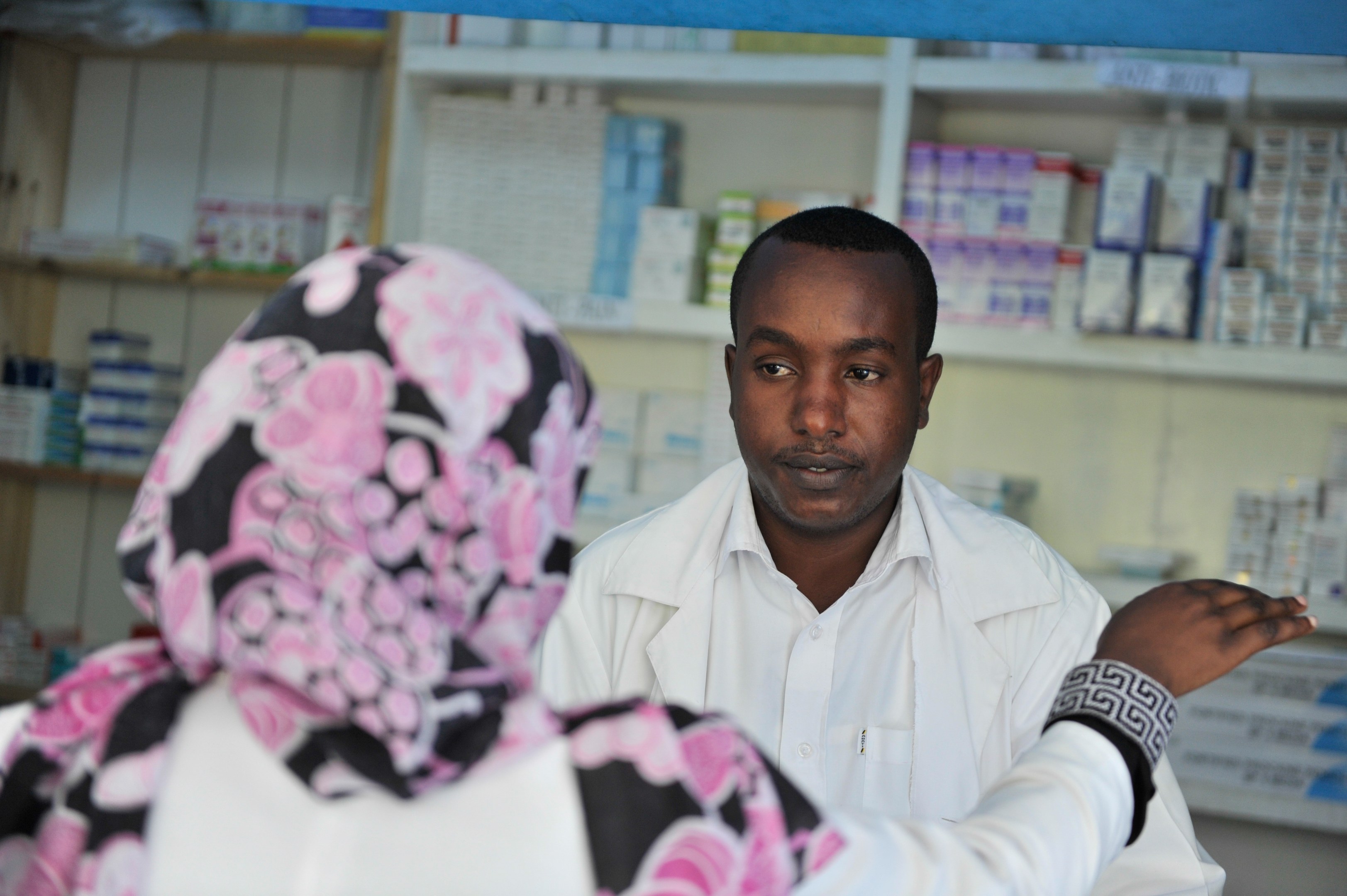 Apothekerin in weißem Kittel, die vor einer sitzenden Frau in einer Apotheke steht und spricht, mit Regalen voller Schachteln und Gegenständen im Hintergrund.