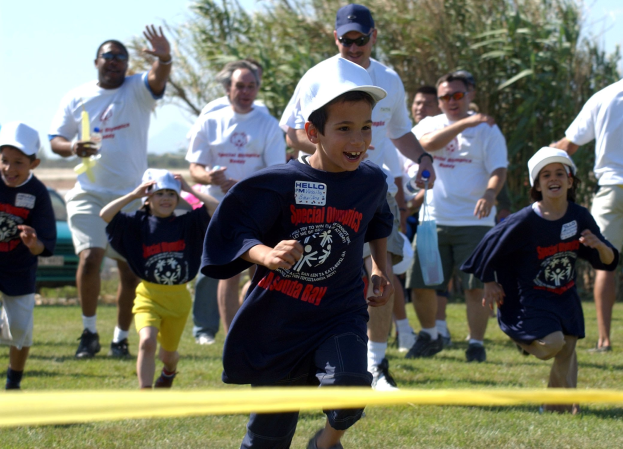Kinder bei einem Lauf auf einer grünen Wiese mit einem gelben Band im Vordergrund, einige tragen Kappen und Schutzbrillen, andere halten Gegenstände, vor einem Hintergrund aus Bäumen und einem klaren blauen Himmel.