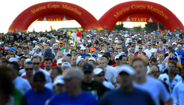 Eine große Gruppe von Läufern bei einem Marathon, mit einem Bogen mit Text im Hintergrund, Bäumen und einem klaren blauen Himmel.