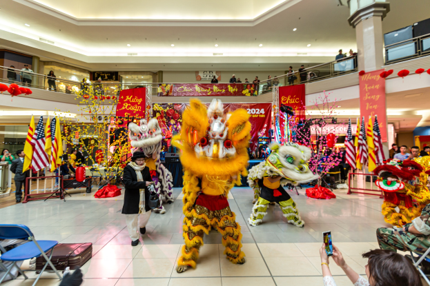Eine lebendige chinesische Neujahrsfeier in einem Einkaufszentrum mit tanzenden Menschen in Kostümen, anderen auf Stühlen sitzend und Mobiltelefone haltend sowie verschiedenen Dekorationen, Flaggen und Bannern im Hintergrund.
