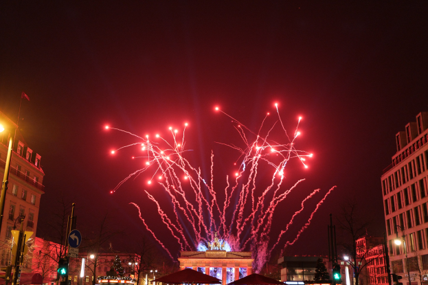 Eine belebte Stadtstraße bei Nacht am Silvesterabend in Berlin, mit Gebäuden, Bäumen, Laternenmasten, Ampeln, Schildern, Zelten, Menschen und einem prächtigen Feuerwerk am Himmel.