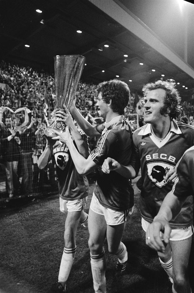 Schwarzes und weißes Foto von Fußballspielern, die mit einem Pokal auf einem Feld feiern, umgeben von einer Menge hinter einem Zaun unter Flutlicht.