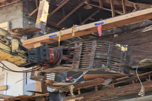 Ein Bagger arbeitet an der Seite eines abbruchreifen Gebäudes, umgeben von Holzplanken und Schutt.