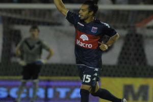 Ein Fussballspieler feiert nach einem Tor auf dem Boden, mit jubelnden Zuschauern und einer Person im Hintergrund, alles vor einem unscharfen, dynamischen Hintergrund.