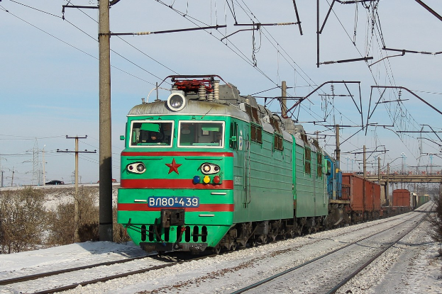Grüne Elektrolokomotive auf schneebedeckten Schienen mit Vegetation, Strommasten mit Drähten, einer Brücke und einem klaren Himmel im Hintergrund.