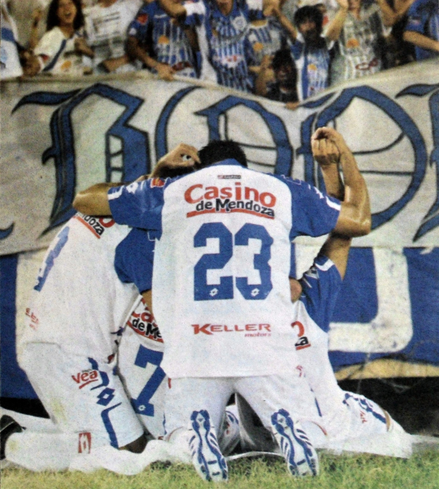 Gruppe von Fußballspielern, die ein Tor auf dem Boden vor einer jubelnden Menge feiern, mit einem Banner mit Text im Hintergrund.