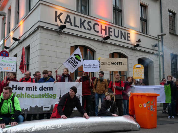 Eine Gruppe von Menschen mit Protestplakaten und Schildern vor einem Gebäude, mit zwei sitzenden Personen im Vordergrund und einem Müllcontainer rechts, vor dem Hintergrund deutscher Stadtarchitektur.