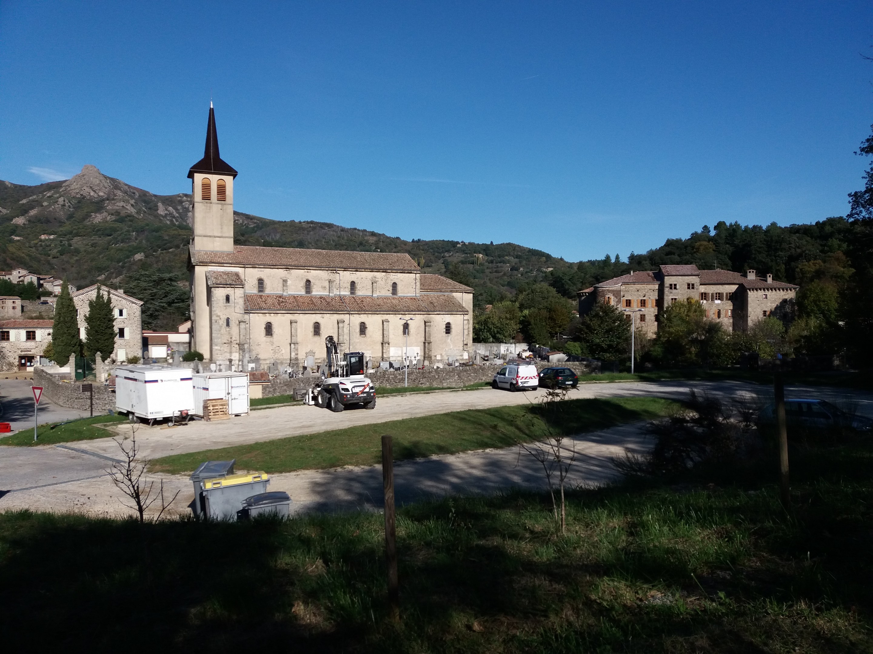 Luftaufnahme einer kleinen Stadt mit einer zentralen Kirche, umgeben von Gebäuden, Bäumen, Straßen, Fahrzeugen und Schildern, vor einem Hügel- und einem klaren blauen Himmel.