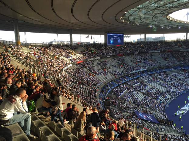 Große Zuschauermenge im Allianz Arena Stadion bei einem Füßtballspiel, mit einer Bühne auf der rechten Seite, Fahnen, Masten, einem Bildschirm und sichtbarem Himmel im Hintergrund.