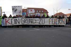 Gruppe von Menschen mit einem Transparent mit der Aufschrift "Kein Grund zu Feiern" während einer Demonstration gegen die Sparpolitik der deutschen Regierung, mit Gebäuden, Bäumen und einem klaren Himmel im Hintergrund.