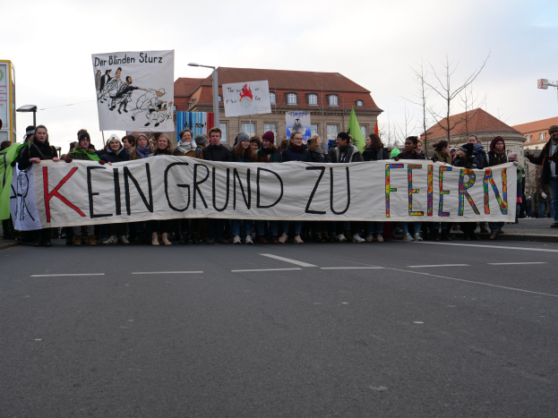 Gruppe von Menschen mit einem Transparent mit der Aufschrift "Kein Grund zu Feiern" während einer Demonstration gegen die Sparpolitik der deutschen Regierung, mit Gebäuden, Bäumen und einem klaren Himmel im Hintergrund.