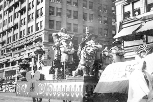 Schwarzes und weißes Foto eines Umzugswagens, der mit Statuen von Menschen und Tieren geschmückt ist und eine von Gebäuden gesäumte Stadtstraße entlangfährt, während Zuschauer von der Straße aus zusehen.