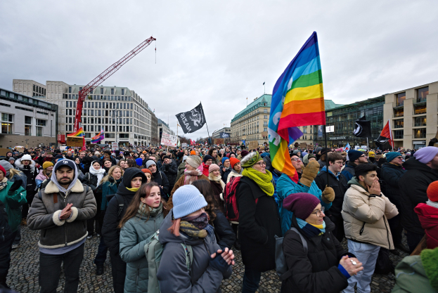 Große Gruppe von Menschen bei einer LGBTQ+-Rechtsdemo in Berlin, die Fahnen und Schilder halten, mit Gebäuden, einem Kran und einem bewölkten Himmel im Hintergrund.
