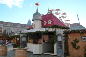 Kleines Gebäude mit rotem Dach und einem Riesenrad davor, umgeben von Festdekorationen und Menschen, mit Gebäuden, Bäumen und wolkenverhangenem Himmel im Hintergrund bei einem Oktoberfest-Event in München, Deutschland.