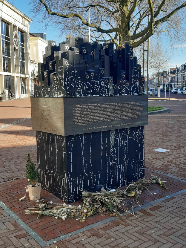 Denkmal in einem Stadtplatz für Holocaust-Opfer, mit einem Baum davor, einer Topfpflanze, verstreuten trockenen Blättern und umliegenden Gebäuden, Fahrzeugen, Pfählen und Bäumen.
