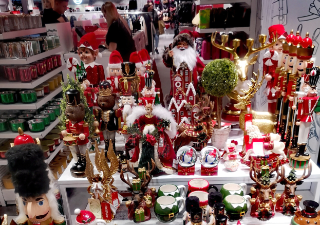 Ladeninneres voller Weihnachtsdekorationen, darunter Spielzeuge, Tassen und andere Gegenst├Ąnde auf Tischen und Regalen, mit Menschen, Lichtern und einer Wand im Hintergrund.