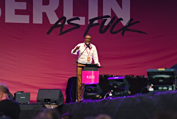 Mann in weißem Hemd und braunen Hosen steht auf einer Bühne und hält ein Mikrofon in der Hand, hinter ihm eine pinkfarbene Fahne mit der Aufschrift "Berlin as Fuck" und Lautsprecher auf der Bühne.