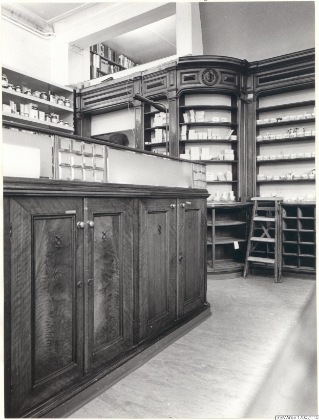 Schwarzes und wei├čes Foto eines vintage Apothekeninneren mit einer Theke, Sch├Ąben, einer Leiter, Regalen mit Beh├Ąltern und Text über dem Bild.