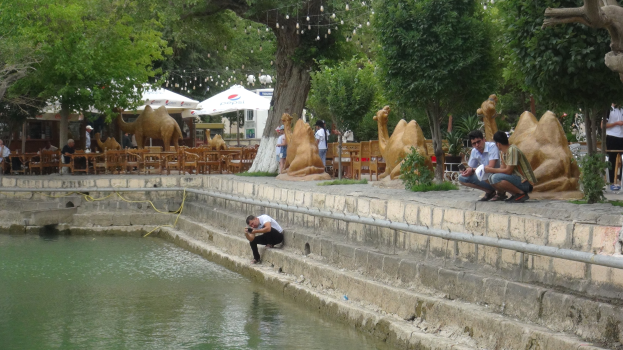 Ein Mann sitzt am Rand eines Teiches umgeben von Menschen, Bäumen, Skulpturen, Möbeln und Sonnenschirmen mit Wasser im Vordergrund und Himmel im Hintergrund.