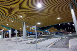 Buswartehäuschen bei Nacht mit Deckenbeleuchtung, Fahrzeuge auf der Straße, Beschilderung, einige Menschen und beleuchtete Gebäude im Hintergrund.