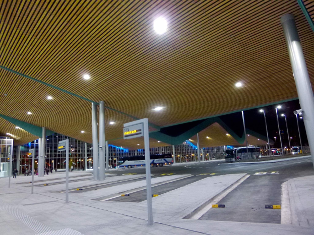 Buswartehäuschen bei Nacht mit Deckenbeleuchtung, Fahrzeuge auf der Straße, Beschilderung, einige Menschen und beleuchtete Gebäude im Hintergrund.