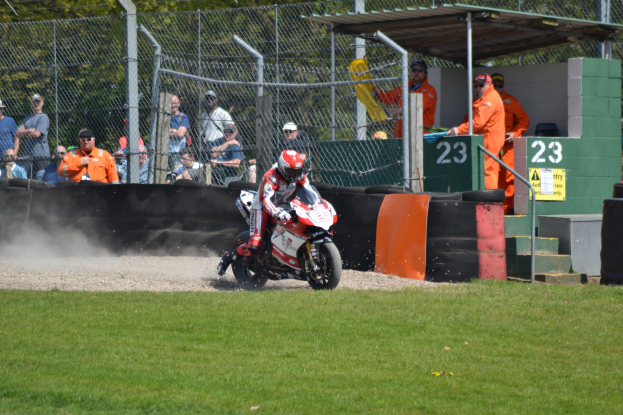 Eine Person in einem Helm und Handschuhen fährt ein Motorrad über ein grasbewachsenes Feld, mit Zuschauern in Mützen und Schutzbrillen, einem eingezäunten Bereich, Treppen mit Geländern und einem nummerierten Schuppen im Hintergrund.