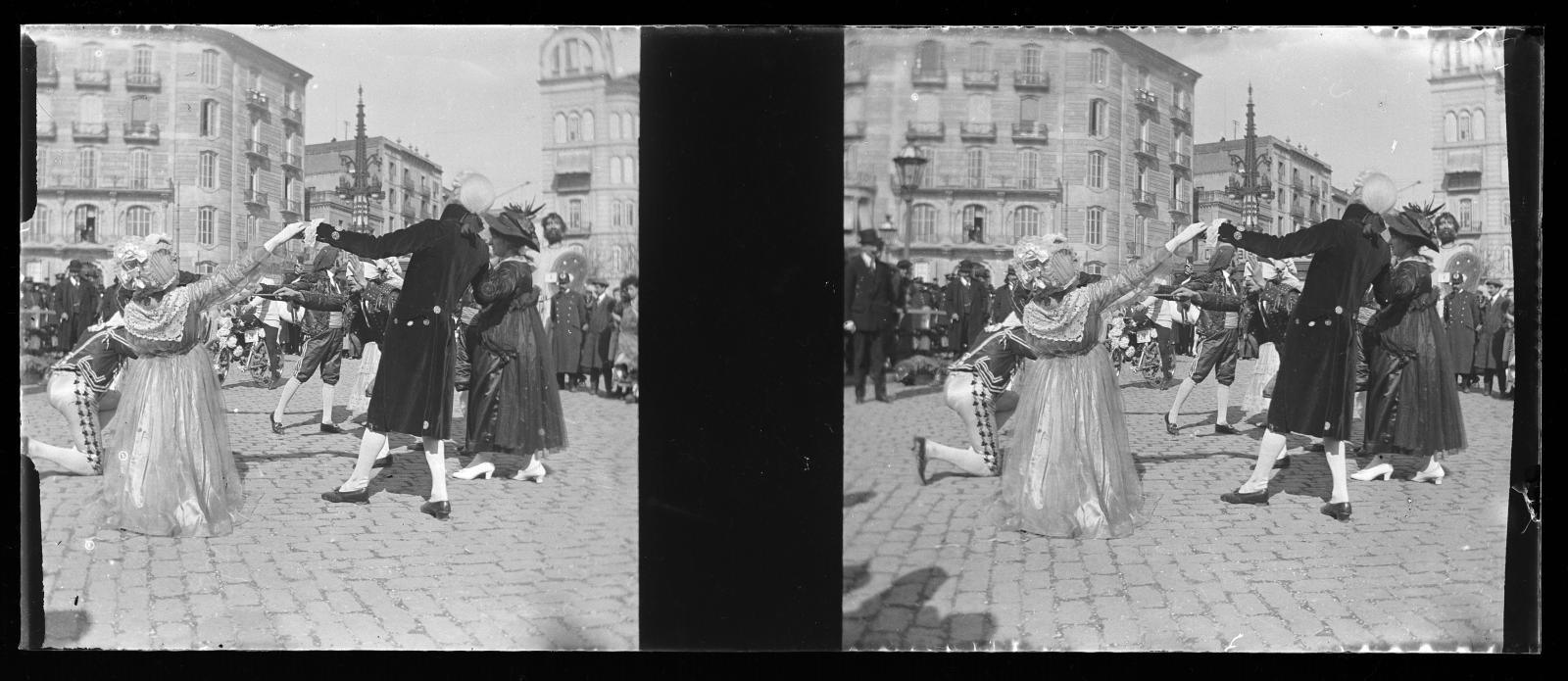 Schwarzes und weißes Foto einer Gruppe von Menschen in Kostümen, die auf der Straße tanzen, umgeben von Gebäuden, Straßenlaternen und Straßenlaternen unter einem klaren Himmel.
