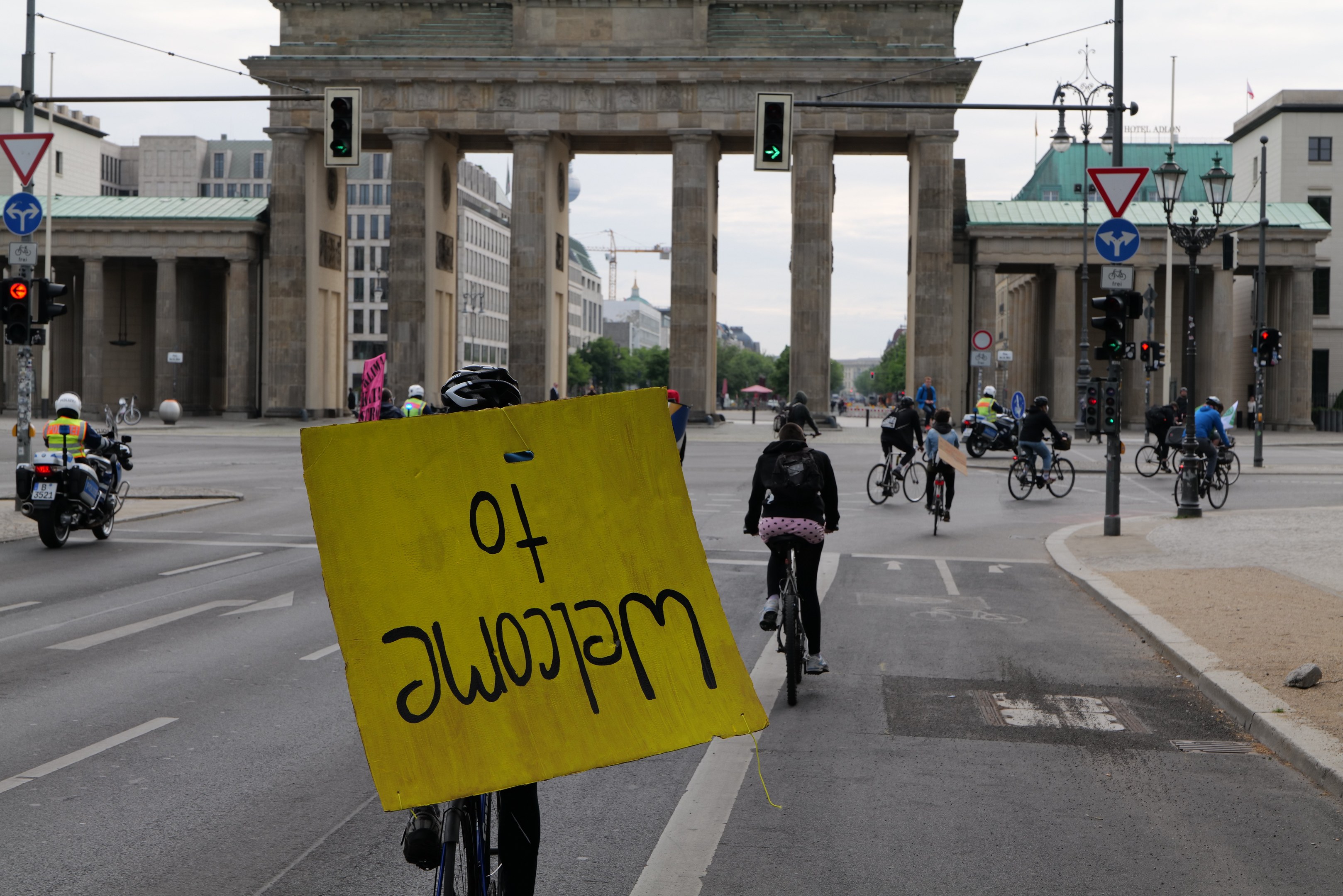 Eine Gruppe von Menschen auf Fahrrädern fährt vor dem Brandenburger Tor in Berlin, Deutschland, vorbei, wobei sie Helme tragen und eine Person ein gelbes Schild hält, mit Laternen, Verkehrszeichen, Gebäuden, Bäumen und einem klaren blauen Himmel im Hintergrund.