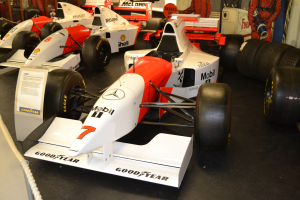 Eine Gruppe von Rennwagen in einem Museum ausgestellt, umgeben von Menschen, ein Schild, eine Seilabsperrung und eine Wand im Hintergrund.
