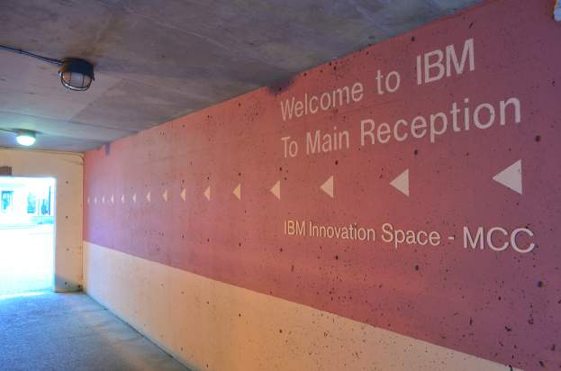 Eine Wand Mit Dem Text "Willkommen Bei IBM" Beleuchtet Von Deckenlichtern, Mit Einer Tür Im Hintergrund Beschriftet "IBM Innovation Space MCC."