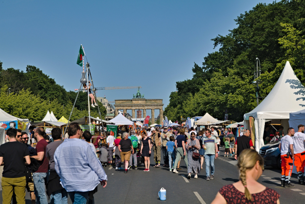 Eine Menge geht eine Straße entlang, gesäumt von Zelten, Fahrzeugen und Bäumen, mit einem Bogen und einem klaren blauen Himmel im Hintergrund und Fahnenmästen auf der linken Seite.
