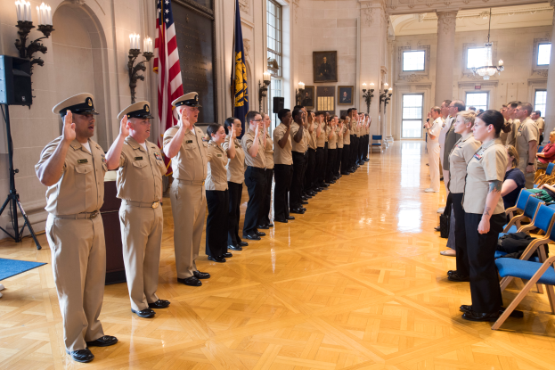 Eine Gruppe uniformierter Personen steht in einem Raum mit Stühlen auf der rechten Seite, Fotorahmen an der Wand, Kerzen, einem Lautsprecher auf der linken Seite und einer Fahne in der Mitte, alle salutierend während einer Militär-Offizier-Zeremonie.